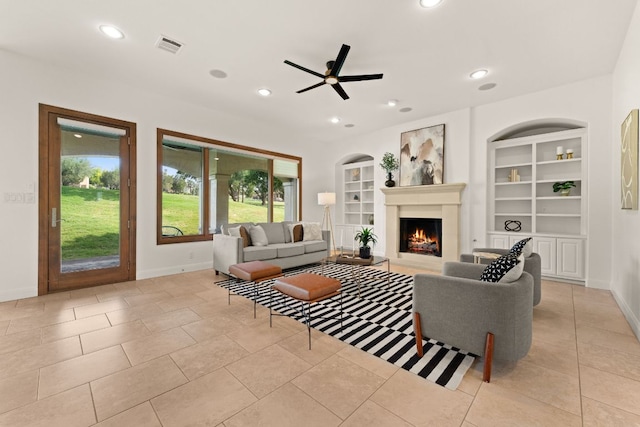 living room featuring baseboards, built in features, ceiling fan, and a lit fireplace