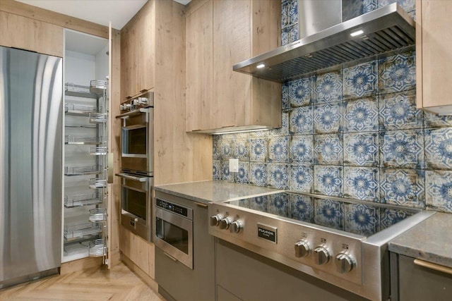 kitchen featuring light brown cabinetry, light stone countertops, wall chimney exhaust hood, stainless steel appliances, and tasteful backsplash