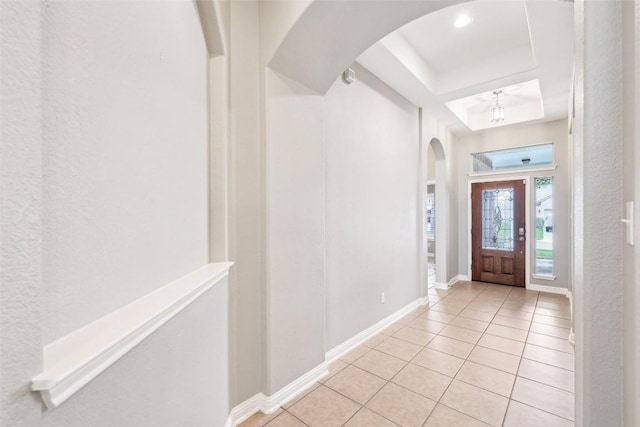 foyer entrance featuring arched walkways, baseboards, a textured wall, a tray ceiling, and light tile patterned flooring