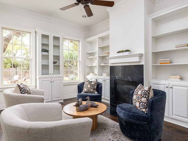 living room featuring a ceiling fan, built in shelves, dark wood-style floors, ornamental molding, and a premium fireplace