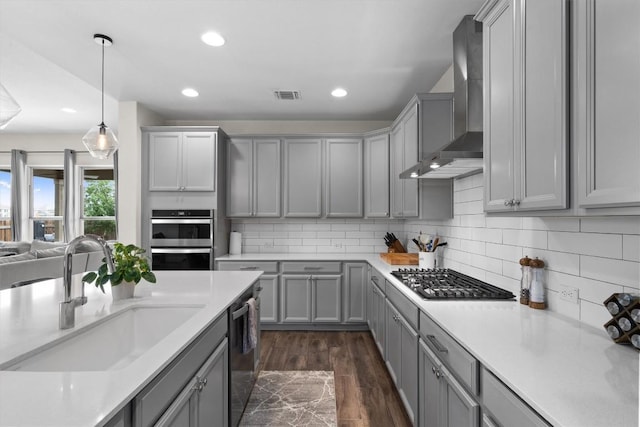 kitchen with appliances with stainless steel finishes, gray cabinetry, a sink, light countertops, and wall chimney exhaust hood
