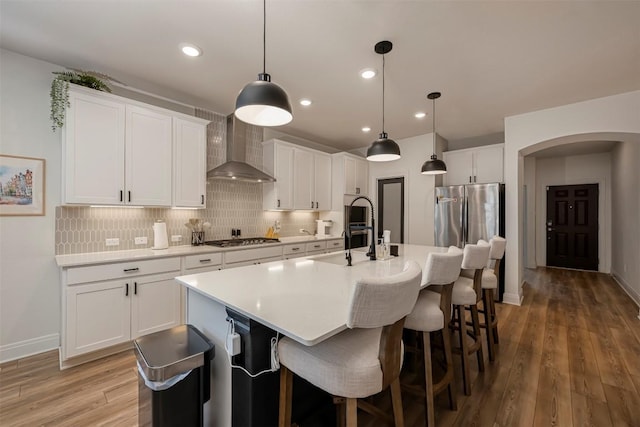 kitchen featuring stainless steel gas cooktop, arched walkways, wall chimney exhaust hood, tasteful backsplash, and a center island with sink