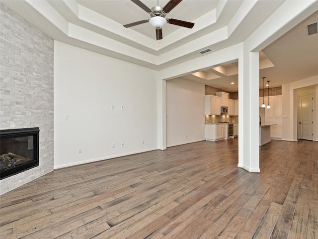 unfurnished living room featuring a fireplace, a ceiling fan, wood finished floors, a tray ceiling, and visible vents