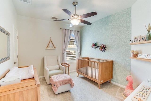 bedroom with light colored carpet, a crib, a ceiling fan, wallpapered walls, and visible vents