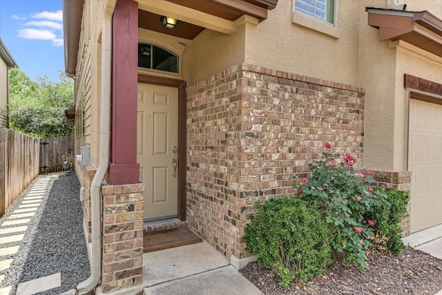 view of exterior entry with stucco siding, brick siding, fence, and a garage
