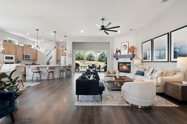 living area featuring dark wood-style flooring, ceiling fan, a brick fireplace, stairs, and recessed lighting