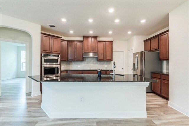 kitchen with a sink, double oven, an island with sink, under cabinet range hood, and visible vents