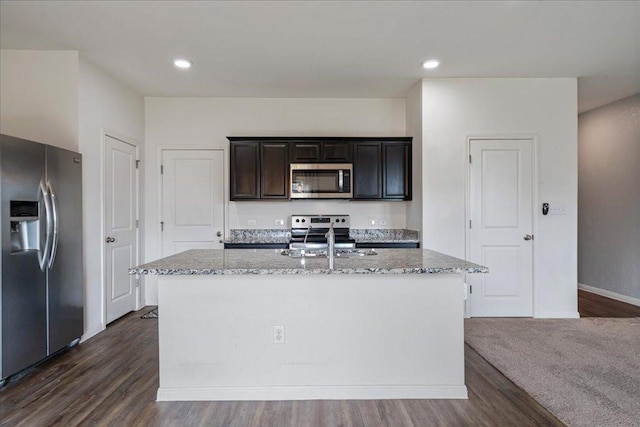 kitchen featuring light stone countertops, appliances with stainless steel finishes, a sink, a kitchen island with sink, and dark wood-type flooring