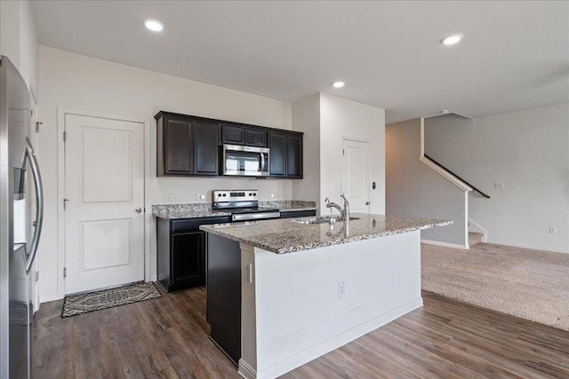 kitchen featuring appliances with stainless steel finishes, light stone counters, a sink, a kitchen island with sink, and dark wood-type flooring