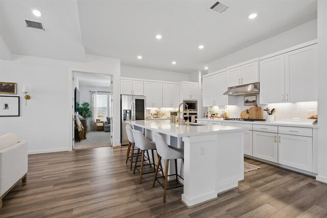 kitchen with appliances with stainless steel finishes, an island with sink, backsplash, under cabinet range hood, and visible vents