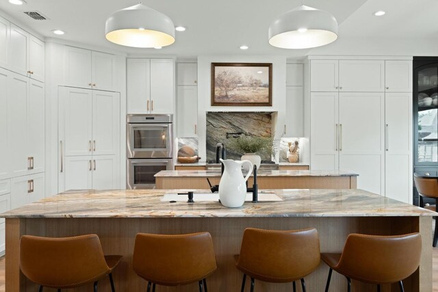 kitchen featuring white cabinets, stainless steel double oven, a center island with sink, and a kitchen bar