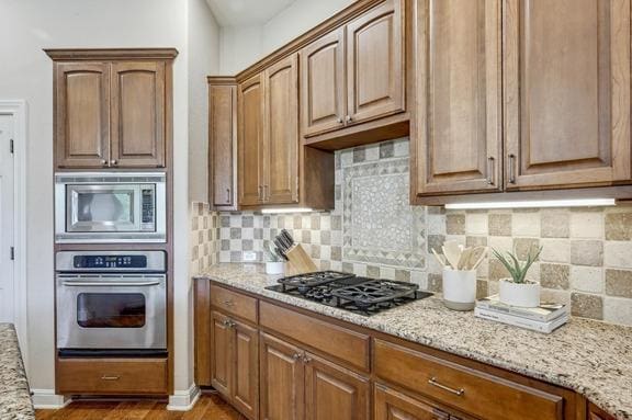 kitchen with decorative backsplash, light wood-style floors, brown cabinets, light stone counters, and appliances with stainless steel finishes