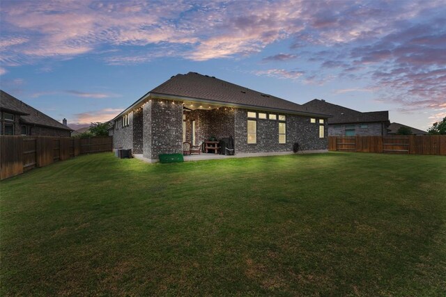 back of property at dusk with a fenced backyard, a yard, central AC, and brick siding