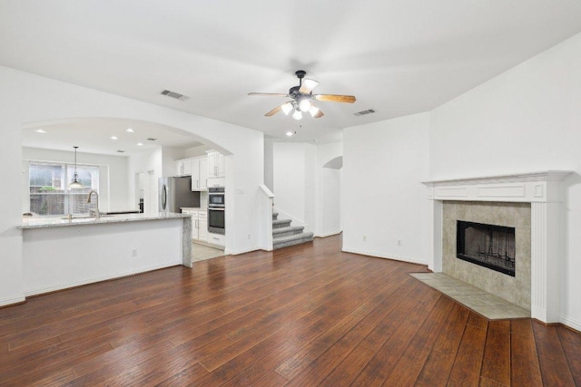 unfurnished living room with ceiling fan, visible vents, a tiled fireplace, stairs, and dark wood-type flooring
