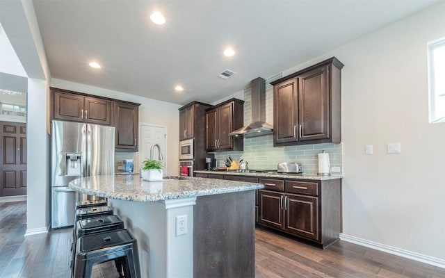 kitchen with dark brown cabinets, appliances with stainless steel finishes, a breakfast bar area, wall chimney exhaust hood, and visible vents