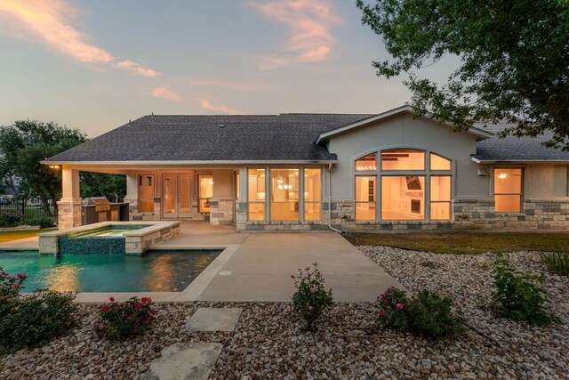 back of property at dusk with roof with shingles, stucco siding, a patio area, and stone siding