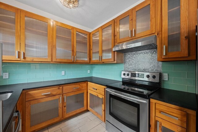 kitchen with under cabinet range hood, stainless steel range with electric cooktop, brown cabinets, and glass insert cabinets