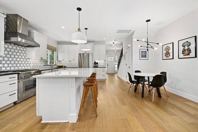 kitchen featuring wall chimney range hood, decorative backsplash, a kitchen island, appliances with stainless steel finishes, and light wood-style flooring