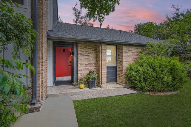 entrance to property featuring a lawn, brick siding, and a shingled roof