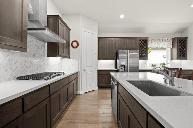 kitchen with dark brown cabinetry, light wood-style floors, wall chimney range hood, a sink, and stainless steel appliances