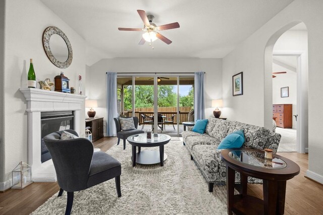 living area with ceiling fan, arched walkways, vaulted ceiling, wood finished floors, and a glass covered fireplace