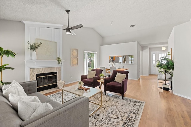living area with a ceiling fan, a textured ceiling, visible vents, a tiled fireplace, and light wood-type flooring