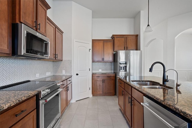 kitchen featuring decorative backsplash, brown cabinetry, appliances with stainless steel finishes, a sink, and dark stone counters
