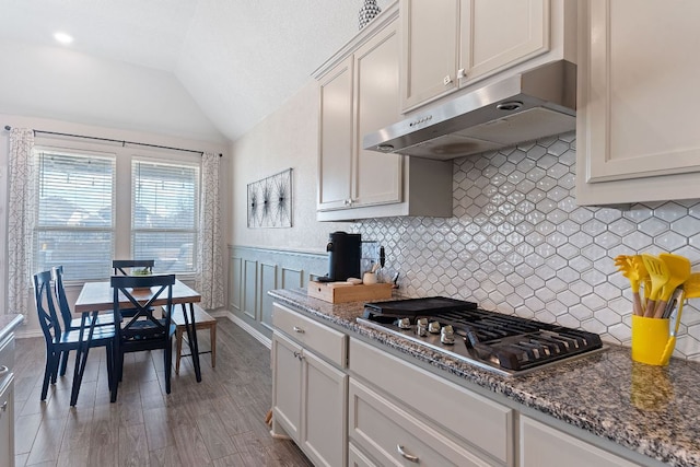 kitchen with dark stone countertops, decorative backsplash, stainless steel gas stovetop, under cabinet range hood, and vaulted ceiling