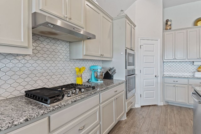 kitchen with decorative backsplash, light stone countertops, under cabinet range hood, stainless steel appliances, and light wood-style flooring