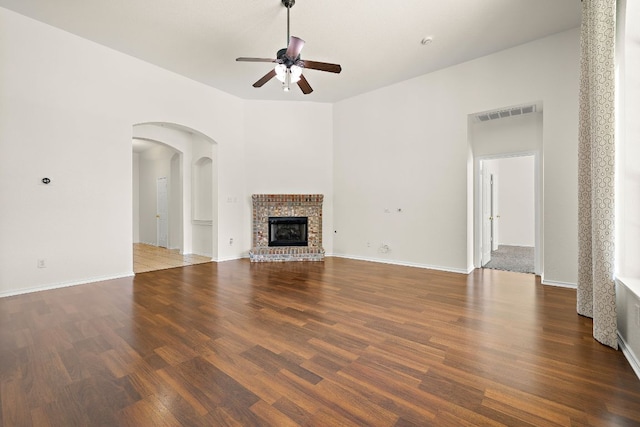unfurnished living room featuring ceiling fan, a brick fireplace, arched walkways, baseboards, and wood finished floors