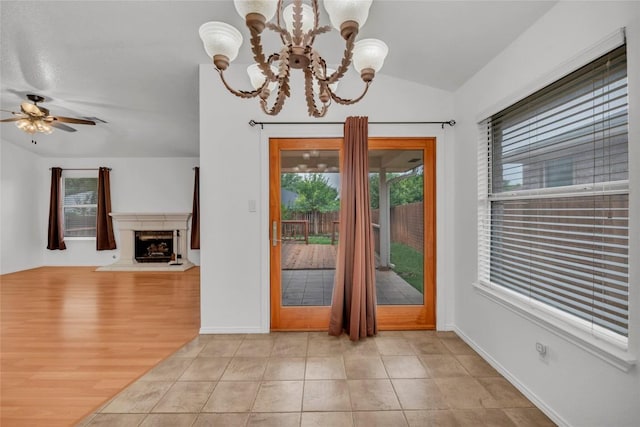 doorway to outside with a fireplace with raised hearth, baseboards, vaulted ceiling, and ceiling fan with notable chandelier