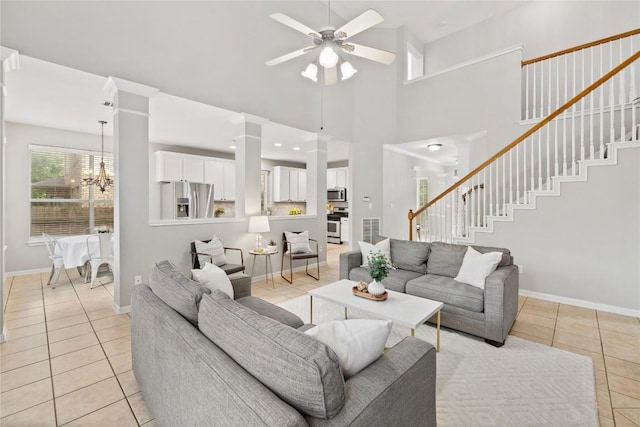 living room featuring light tile patterned flooring, ceiling fan with notable chandelier, baseboards, ornate columns, and stairway