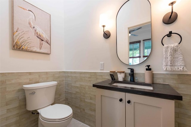 bathroom with a ceiling fan, tile walls, wainscoting, vanity, and toilet
