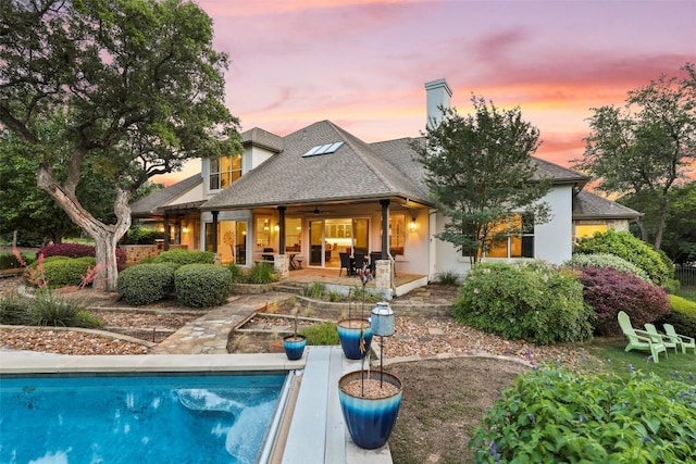 back of property at dusk featuring a shingled roof, a patio area, an outdoor pool, and a chimney