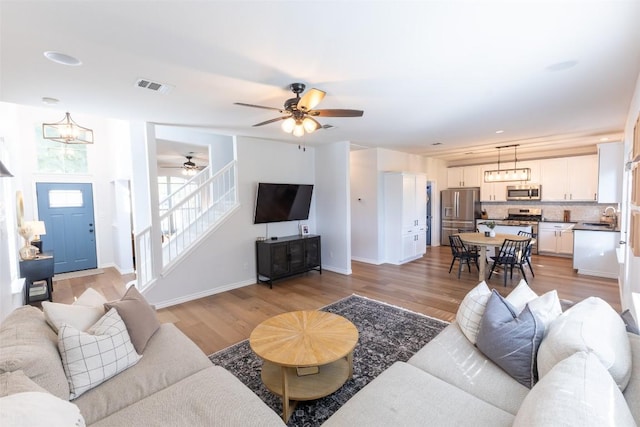 living area with visible vents, stairs, baseboards, light wood finished floors, and ceiling fan with notable chandelier