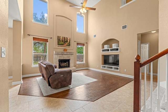 tiled living area featuring a high ceiling, ceiling fan, a stone fireplace, baseboards, and visible vents