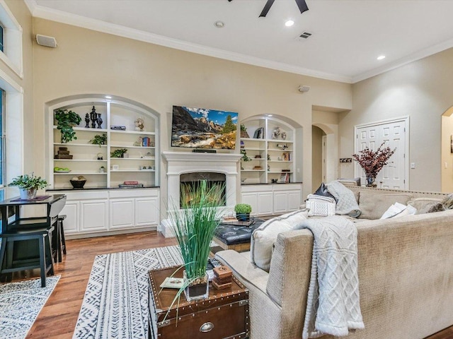 living area with crown molding, light wood-style floors, a fireplace with raised hearth, built in shelves, and a ceiling fan