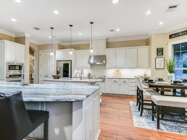 5709 Laguna Cliff Ln, Austin, TX 78734 - kitchen with appliances with stainless steel finishes, under cabinet range hood, a kitchen island with sink, white cabinetry, and ornamental molding