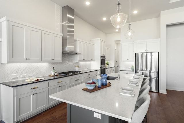 kitchen with wall chimney range hood, decorative backsplash, a sink, stainless steel appliances, and an island with sink