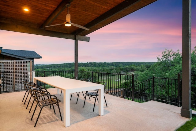 patio terrace at dusk with ceiling fan, a wooded view, and a balcony