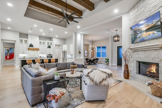 living room with a high ceiling, beam ceiling, a fireplace, ceiling fan with notable chandelier, and light wood-type flooring