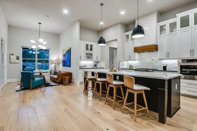 kitchen with visible vents, stainless steel oven, a center island with sink, light wood finished floors, and backsplash