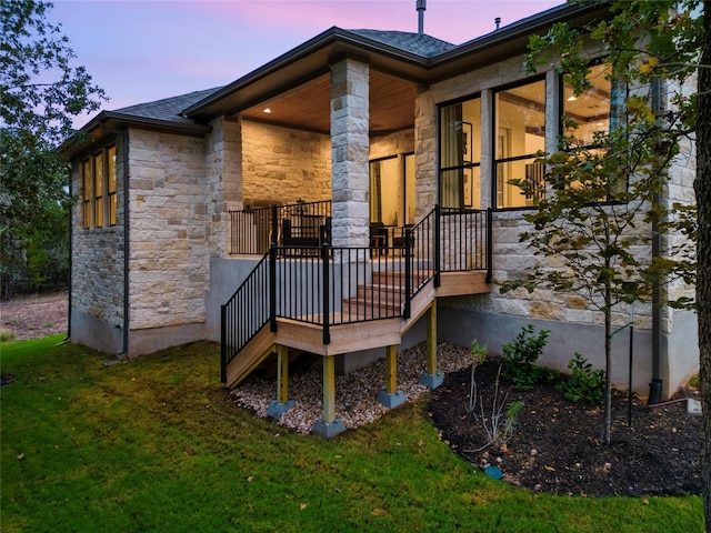 back of house at dusk featuring a lawn, roof with shingles, and stone siding