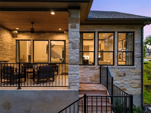 exterior entry at dusk featuring ceiling fan, a shingled roof, and stone siding