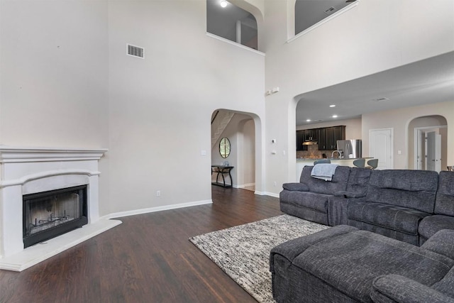 living room featuring arched walkways, dark wood-style floors, a towering ceiling, a fireplace with raised hearth, and baseboards