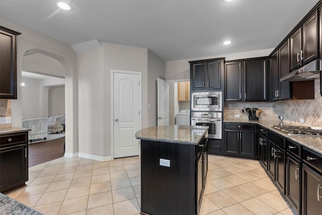 kitchen with appliances with stainless steel finishes, under cabinet range hood, decorative backsplash, light stone countertops, and arched walkways