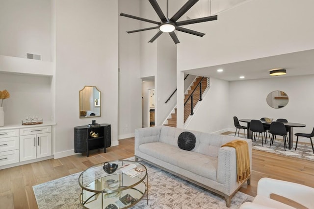 living room featuring a high ceiling, ceiling fan, light wood-style flooring, stairs, and visible vents