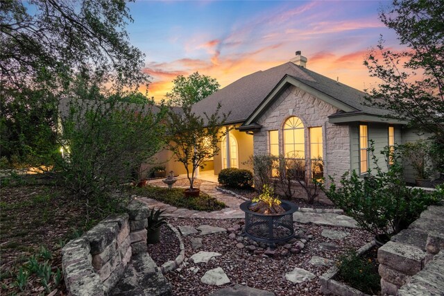 back of property at dusk with a chimney, an outdoor fire pit, roof with shingles, and stone siding