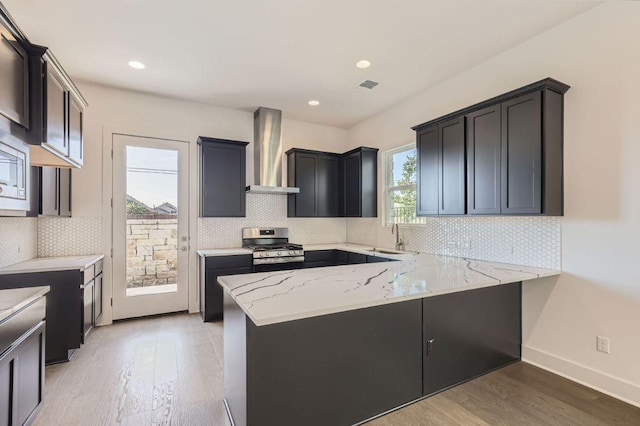 kitchen featuring a peninsula, light stone countertops, wall chimney range hood, stainless steel range with gas stovetop, and light wood finished floors
