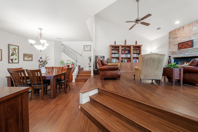 living room with visible vents, a brick fireplace, wood finished floors, stairway, and ceiling fan with notable chandelier